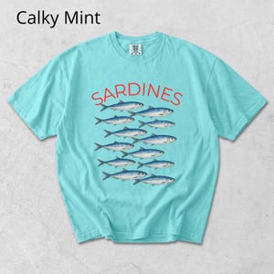 May include: A turquoise t-shirt featuring the word "SARDINES" in red, above a graphic of fish. The shirt has a crew neck and short sleeves. The text "Calky Mint" is in the upper left corner of the shirt.
