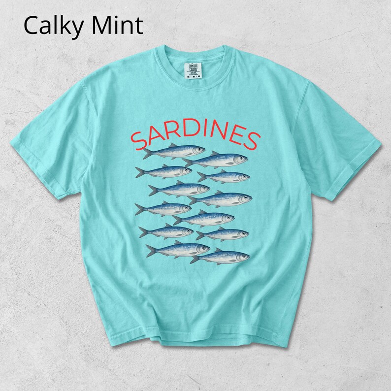May include: A turquoise t-shirt featuring the word "SARDINES" in red, above a graphic of fish. The shirt has a crew neck and short sleeves. The text "Calky Mint" is in the upper left corner of the shirt.