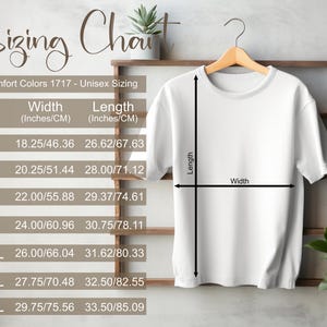 May include: White t-shirt hanging on a wooden rack, displaying a sizing chart. The chart includes measurements in inches and centimetres for sizes S through 4XL. The text reads "Sizing Chart" and "Comfort Colors 1717 - Unisex Sizing."