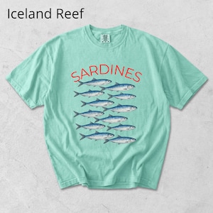 May include: A pale green t-shirt featuring the word "SARDINES" in red above a graphic of blue fish. The text "Iceland Reef" is in the upper left corner. The shirt has a crew neck and short sleeves.