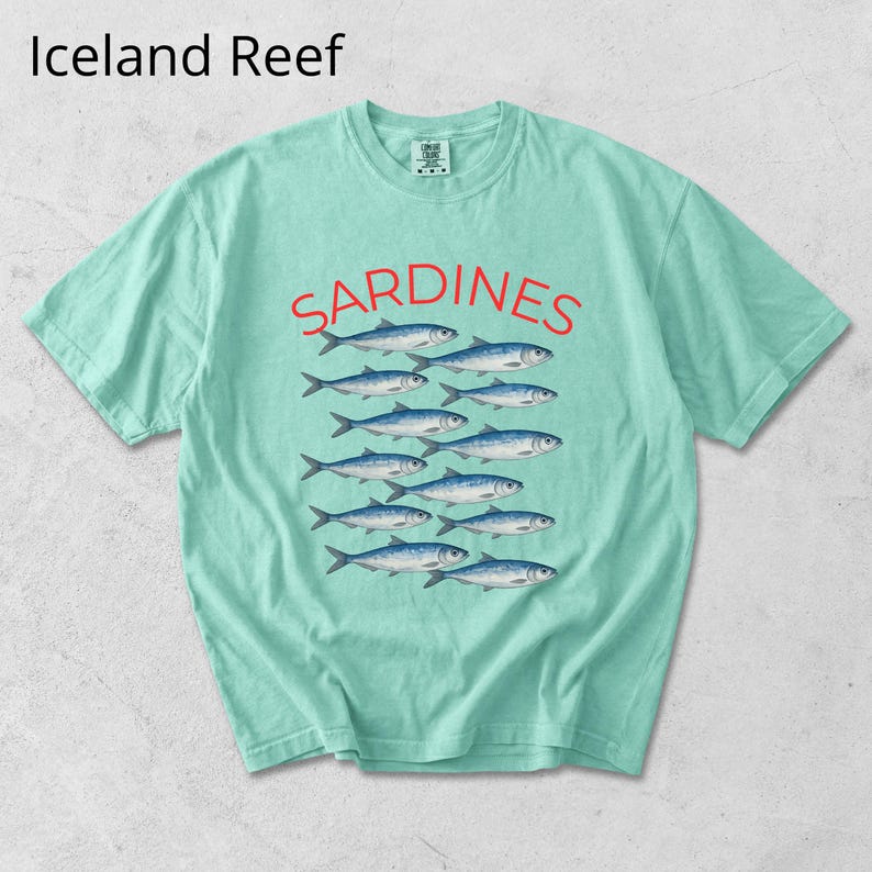May include: A pale green t-shirt featuring the word "SARDINES" in red above a graphic of blue fish. The text "Iceland Reef" is in the upper left corner. The shirt has a crew neck and short sleeves.