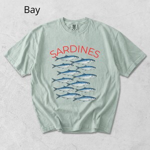 May include: A pale green t-shirt featuring the word "SARDINES" in red, above a graphic of blue fish. The word "Bay" is in the upper left corner. The shirt has a crew neck and short sleeves.