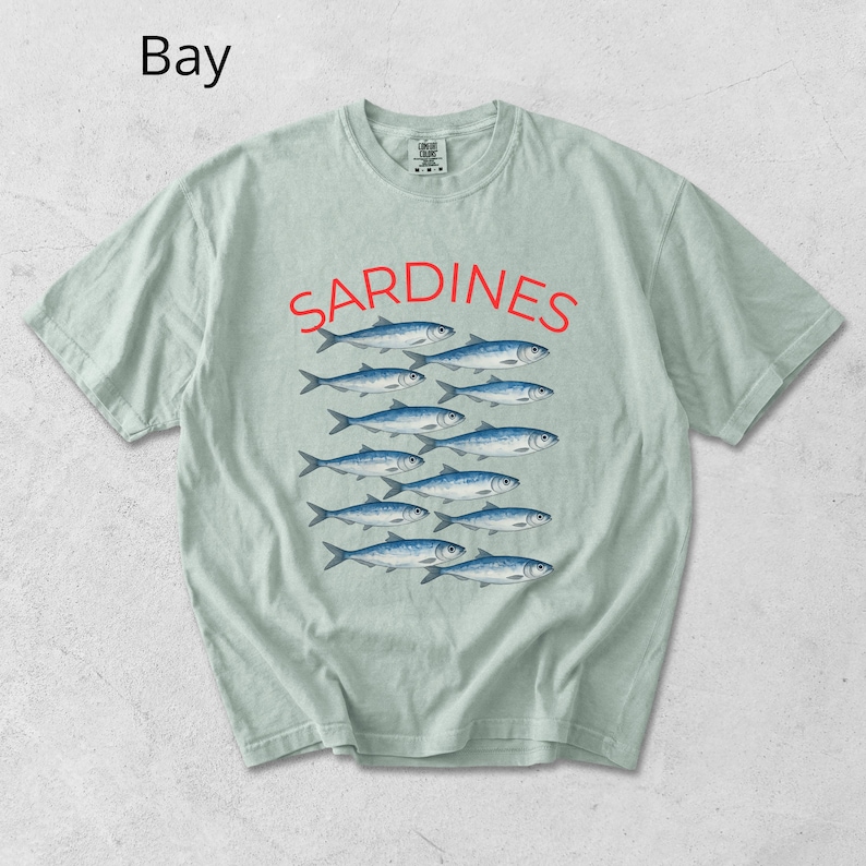 May include: A pale green t-shirt featuring the word "SARDINES" in red, above a graphic of blue fish. The word "Bay" is in the upper left corner. The shirt has a crew neck and short sleeves.