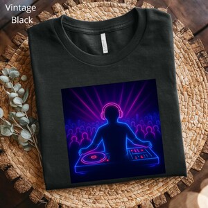 May include: Black t-shirt featuring a neon graphic of a DJ performing at a concert. The graphic shows a DJ with headphones, a turntable, and a crowd, with pink and blue neon lights. The text "Vintage Black" is in the upper left corner.