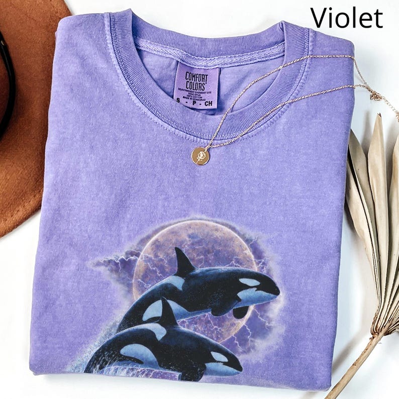 May include: A violet Comfort Colors t-shirt with a graphic of two orcas leaping in front of a full moon and lightning. A gold necklace with a small pendant is on the shirt. The shirt is folded and displayed on a white surface.