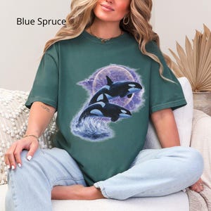 May include: Teal t-shirt with a graphic of two orcas leaping from water, set against a purple moon and lightning. The text "Blue Spruce" is printed at the top left. The shirt has short sleeves and a relaxed fit.