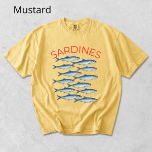May include: Mustard-coloured t-shirt featuring the word "SARDINES" in red, above a pattern of blue fish. The shirt has a crew neck and short sleeves, suitable for everyday wear.