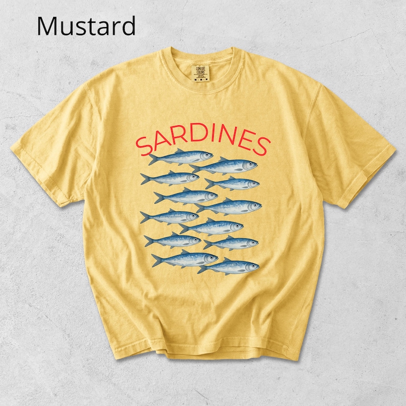 May include: Mustard-coloured t-shirt featuring the word "SARDINES" in red, above a pattern of blue fish. The shirt has a crew neck and short sleeves, suitable for everyday wear.