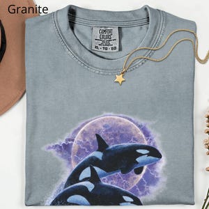 May include: A folded, light grey Comfort Colors t-shirt featuring a graphic of two orcas leaping before a purple moon and lightning. A gold star pendant necklace rests on the shirt. The word "Granite" is in the upper left corner.