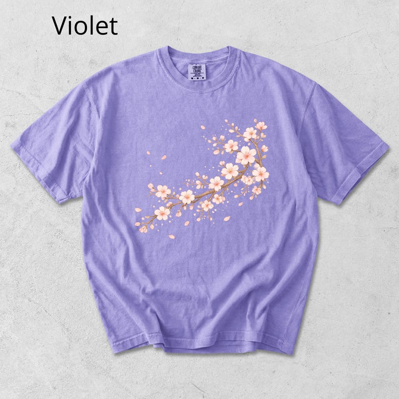 May include: Violet-colored t-shirt featuring a floral branch design with white and peach blossoms. The short-sleeved shirt has a crew neck and a small tag near the collar. The word "Violet" is printed in the upper left corner.