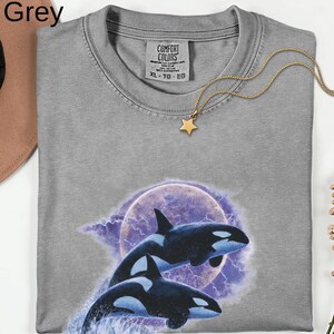 May include: A grey Comfort Colors t-shirt with a graphic of two orcas against a purple moon and lightning. The shirt has a crew neck and short sleeves. A gold star pendant necklace is draped across the shirt.