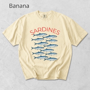 May include: A pale yellow t-shirt featuring the word "SARDINES" in red, above a graphic of blue fish. The shirt has a crew neck and short sleeves. The word "Banana" is in the top left corner.