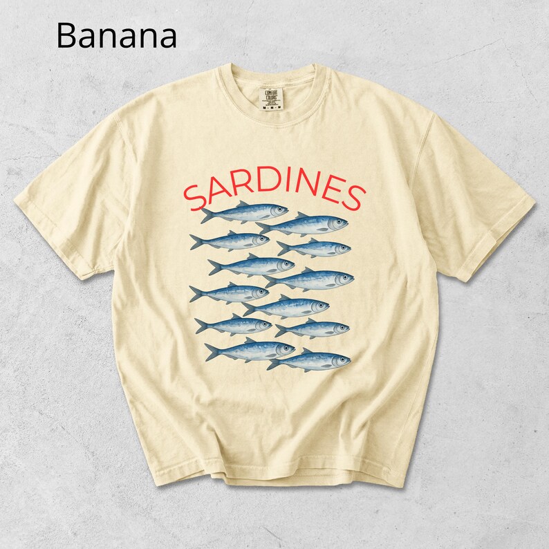May include: A pale yellow t-shirt featuring the word "SARDINES" in red, above a graphic of blue fish. The shirt has a crew neck and short sleeves. The word "Banana" is in the top left corner.