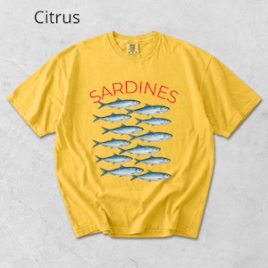 May include: Mustard yellow t-shirt featuring the word "SARDINES" in red, above a pattern of blue fish. The shirt has a crew neck and short sleeves. The word "Citrus" is in the upper left corner.