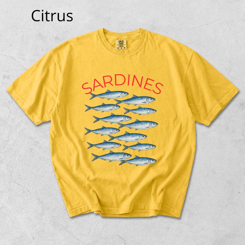 May include: Mustard yellow t-shirt featuring the word "SARDINES" in red, above a pattern of blue fish. The shirt has a crew neck and short sleeves. The word "Citrus" is in the upper left corner.