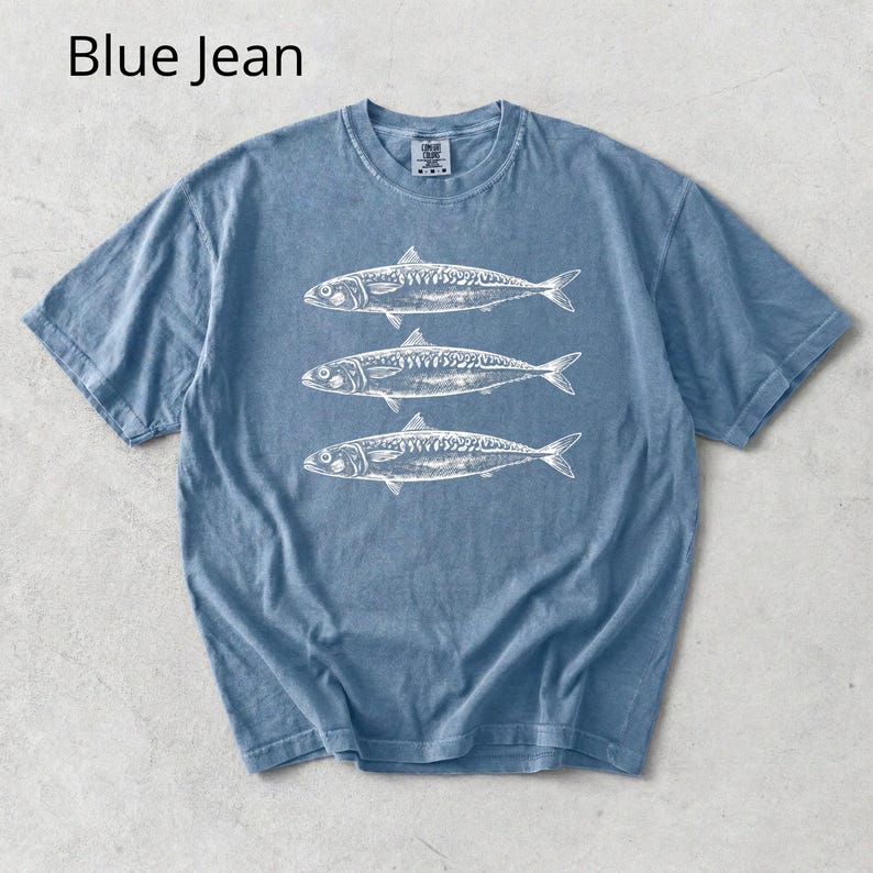 May include: A blue jean colored t-shirt featuring a white illustration of three fish. The t-shirt has short sleeves and a crew neck. The text "Blue Jean" is in the upper left corner.