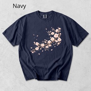 May include: Navy blue t-shirt featuring a floral branch design with pink blossoms and falling petals. The word "Navy" is printed in white at the top left. The t-shirt has a crew neck and short sleeves.