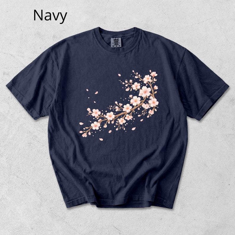 May include: Navy blue t-shirt featuring a floral branch design with pink blossoms and falling petals. The word "Navy" is printed in white at the top left. The t-shirt has a crew neck and short sleeves.