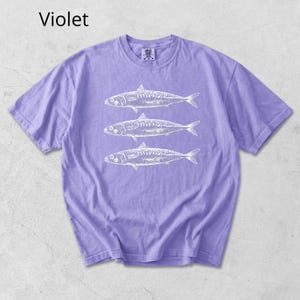 May include: A violet-colored t-shirt featuring a white graphic of three fish. The text "Violet" is in the upper left corner. The shirt has short sleeves and a crew neck.