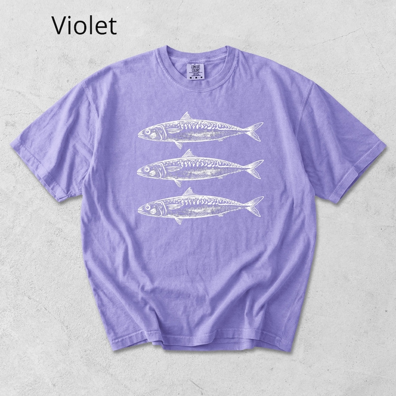 May include: A violet-colored t-shirt featuring a white graphic of three fish. The text "Violet" is in the upper left corner. The shirt has short sleeves and a crew neck.