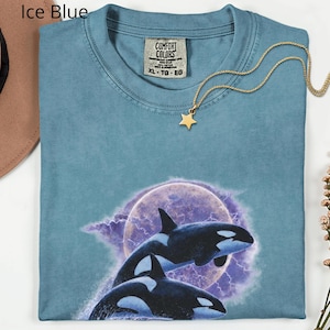 May include: Ice blue Comfort Colors t-shirt with a graphic of two orcas leaping before a purple moon and lightning. A gold star pendant hangs from a delicate chain. The shirt is size XL-TG-EG.