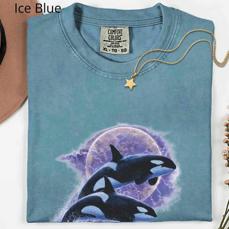 May include: Ice blue Comfort Colors t-shirt with a graphic of two orcas leaping before a purple moon and lightning. A gold star pendant hangs from a delicate chain. The shirt is size XL-TG-EG.