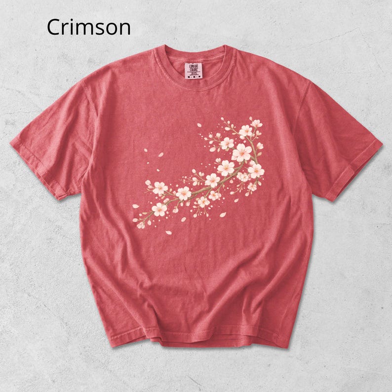 May include: Crimson-colored t-shirt featuring a floral branch design with white and light pink blossoms. The short-sleeved shirt has a crew neck and a slightly faded appearance. The word "Crimson" is printed in the upper left corner.