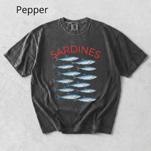 May include: Dark grey t-shirt featuring the word "SARDINES" in red above a row of blue fish. The shirt has a vintage, washed appearance. The word "Pepper" is in the top left corner.