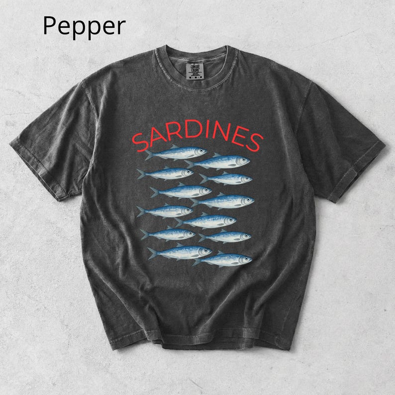 May include: Dark grey t-shirt featuring the word "SARDINES" in red above a row of blue fish. The shirt has a vintage, washed appearance. The word "Pepper" is in the top left corner.