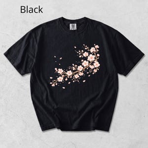 May include: Black t-shirt featuring a floral branch design with pink blossoms and falling petals. The text "Black" is visible in the upper left corner. This short-sleeved tee is a comfortable and stylish addition to any wardrobe.