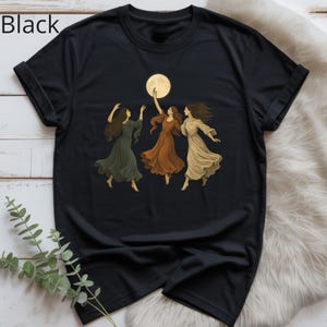 Pode incluir: T-shirt preta com um desenho de três mulheres dançando sob a lua cheia. As mulheres usam vestidos esvoaçantes em tons de verde, laranja e creme. A palavra "Black" está impressa no canto superior esquerdo.