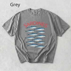 May include: Grey t-shirt featuring the word "SARDINES" in red, above a pattern of blue fish. The shirt has a crew neck and short sleeves. The word "Grey" is in the top left corner.