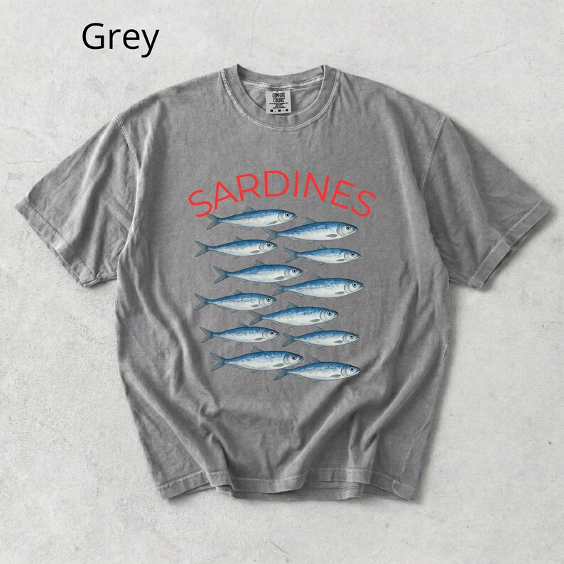 May include: Grey t-shirt featuring the word "SARDINES" in red, above a pattern of blue fish. The shirt has a crew neck and short sleeves. The word "Grey" is in the top left corner.