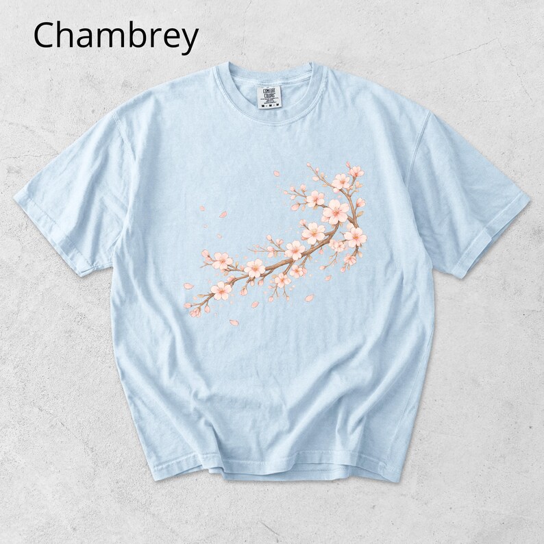 May include: Light blue t-shirt with a floral branch design featuring pink blossoms and falling petals. The word "Chambrey" is printed at the top left. The shirt has short sleeves and a crew neck.