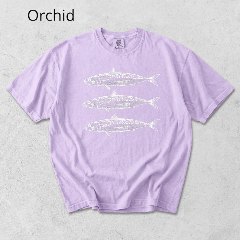 May include: Orchid-colored t-shirt with a crew neck and short sleeves. The shirt features a white graphic of three fish. The word "Orchid" is printed in the upper left corner of the image.
