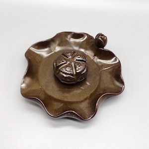 May include: A brown ceramic incense burner shaped like a lotus leaf with a small flower bud on the side. The burner has a round, brown incense holder in the center.