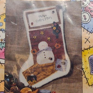 May include: A brown and white felt Christmas stocking with a snowman, stars, and the text "Merry Christmas".