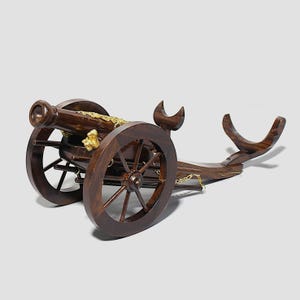 Op de afbeelding: Een houten kanonmodel met een donkerbruine afwerking. Het kanon heeft twee grote houten wielen, een decoratief goudkleurig accent en een gebogen steunstructuur. Een kleine gouden ketting verbindt het kanon met de steun.