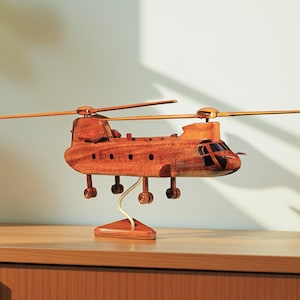 May include: A detailed wooden model of a Chinook helicopter, featuring rotating blades and intricate design. The model is crafted from a warm-toned wood and stands on a wooden base with a white support. The helicopter model is a decorative item.