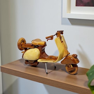 May include: A wooden scooter model with a light brown and dark brown finish. The scooter has a rounded front, a seat, and a rear wheel. The model is displayed on a wooden shelf. The scooter is approximately 10 inches long.
