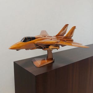 May include: A wooden model of a fighter jet, showcasing a detailed design with a glossy, polished finish. The jet is a warm, medium brown color, mounted on a matching wooden stand. The cockpit is black, and the wings are angled upwards.