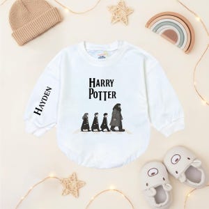 Puede incluir: Body blanco de bebé con mangas largas con el texto "Harry Potter" y un gráfico de personajes caminando. La manga tiene el nombre "Hayden" impreso verticalmente. También se ve un gorro de punto beige y decoraciones en forma de estrella.