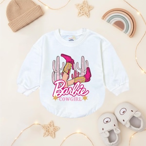 May include: White long-sleeved baby bodysuit with a graphic of pink cowboy boots, cacti, and the text "Barbie Cowgirl". The design includes gold stars and a pink Barbie logo. The bodysuit is surrounded by baby accessories.