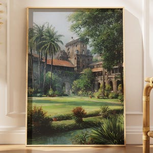 May include: A framed landscape painting depicts a lush green lawn with a pond, palm trees, and a large building with a red tile roof. The artwork is in a gold frame and leans against a white wall.
