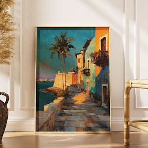 Op de afbeelding: Een ingelijste kunstdruk die een kustscène weergeeft met gebouwen in tinten oranje, blauwgroen en blauw. Een palmboom staat in de buurt van de gebouwen en een stenen voetpad leidt naar het water. Het kunstwerk wordt op een witte muur getoond.