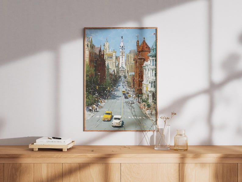 May include: A framed painting depicts a city street scene with buildings lining both sides. Two vintage cars, one yellow and one white, drive down the center of the road. The artwork is in a wooden frame and hangs on a white wall.
