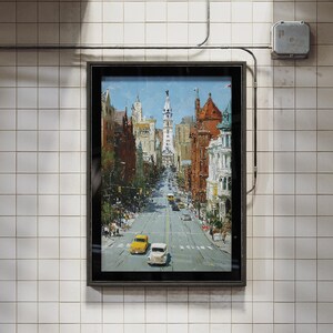 May include: A framed painting of a city street scene, featuring buildings in shades of brown, red, and white. The artwork depicts a long road with cars and trees, under a blue sky. The frame is black, and the background is a tiled wall.