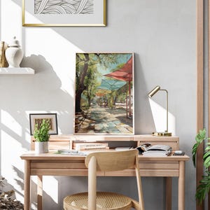 Puede incluir: Una pintura enmarcada de un paisaje de un paseo sombreado con toldos coloridos. La obra de arte se exhibe en un escritorio de madera con una peque&ntilde;a planta en maceta, una l&aacute;mpara de escritorio y una silla de madera. Una estanter&iacute;a decorativa y una obra de arte abstracta enmarcada son visibles.