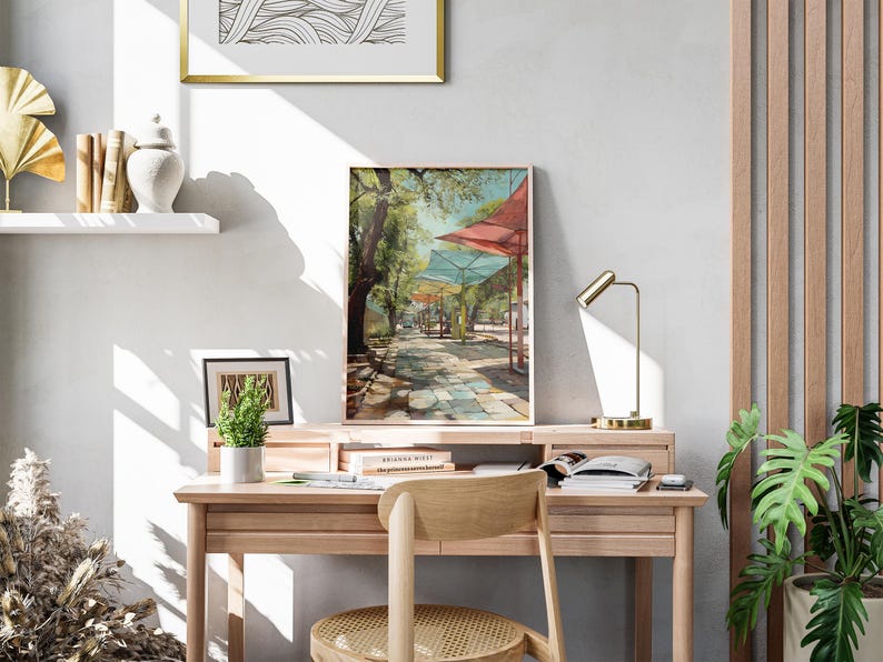 Puede incluir: Una pintura enmarcada de un paisaje de un paseo sombreado con toldos coloridos. La obra de arte se exhibe en un escritorio de madera con una peque&ntilde;a planta en maceta, una l&aacute;mpara de escritorio y una silla de madera. Una estanter&iacute;a decorativa y una obra de arte abstracta enmarcada son visibles.