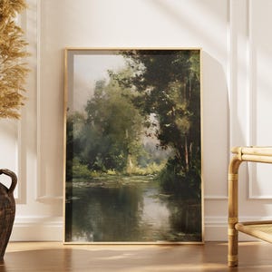 Puede incluir: Una pintura de paisaje enmarcada representa una serena escena fluvial con exuberantes árboles y follaje verde. La obra de arte se exhibe sobre un suelo de madera, junto a un jarrón decorativo con plantas secas. El marco es dorado.